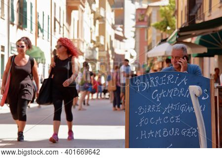 Lerici, Italy - Summer2020: Tourists Stroll The Central Street And A Man And Sit Behind The Menu Of 