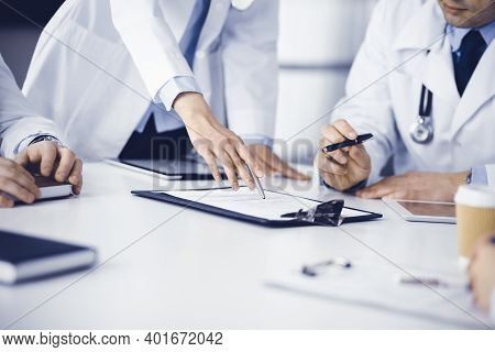 Group Of Unknown Doctors Are Sitting At The Desk And Discussing Medical Treatment, Using A Clipboard