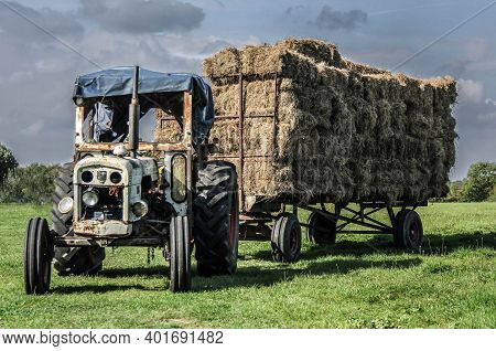 Old Neglected Blue Tractor Pulling A Triler Of Hay In A Field At Harvest Time In Summer