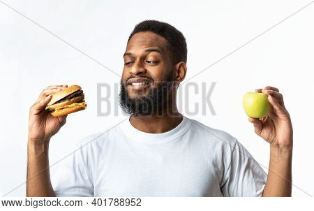 Healthy Vs Unhealthy Food. Hungry African Man Choosing Between Burger And Apple Standing In Studio O