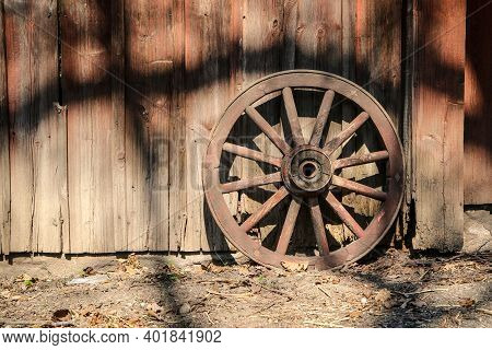 The Old Wooden Wheel From A Cart Is Standing By The House Fith Facade From The Planks.