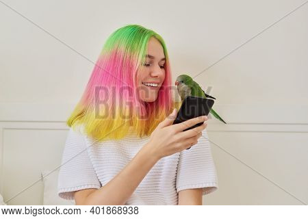 Trendy Teen Girl With Smartphone And Young Green Quaker Parrot On Her Shoulder, At Home On Bed. Pet 