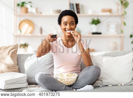 Millennial Black Lady With Popcorn Switching Channels On Tv, Sitting On Sofa At Home. African Americ