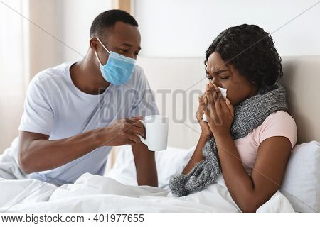 Black Man In Protective Face Mask Taking Care Of His Sick Wife, Giving Hot Drink, Bedroom Interior, 