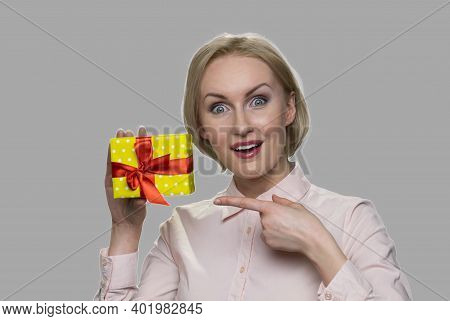Happy Shocked Woman Pointing At Gift Box. Excited Woman Holding Peresent Box On Gray Background.
