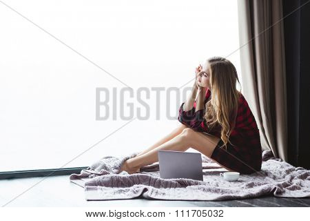 Beautiful pensive young woman with long hair in checkered shirt sitting on coverlet on the floor with laptop and cup of coffee