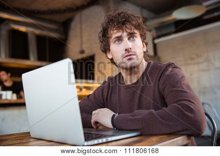 Thoughtful curly pensive concentrated handsome young man in brown sweetshirt working using laptop in cafe