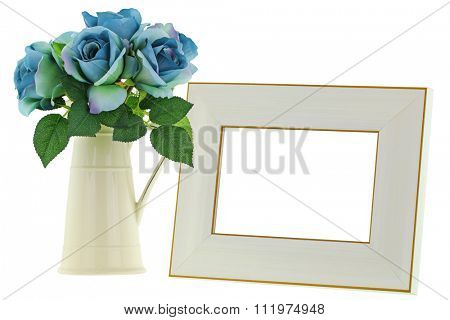 A yellow vintage enamel ceramic jug vase with blue green roses next to blank beige wooden picture frame with golden border, isolated on white background
