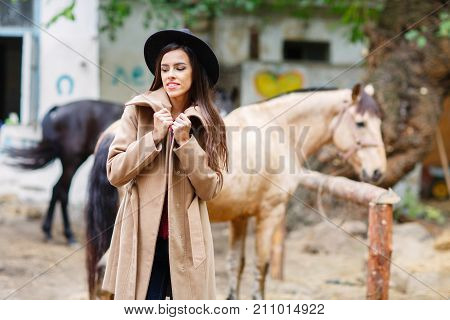 A white brunette girl in a black hat and a light coat stands and smiles near two horses. Outside.