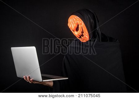Anonymous male hacker with pumpkin halloween mask carrying laptop computer. Internet security and cyber attack concepts.