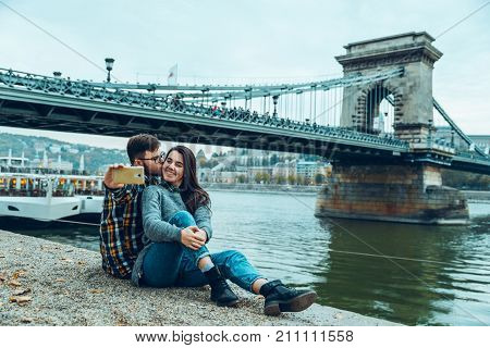 lovely cople sitting on the beach and taking selfie old bridge on background