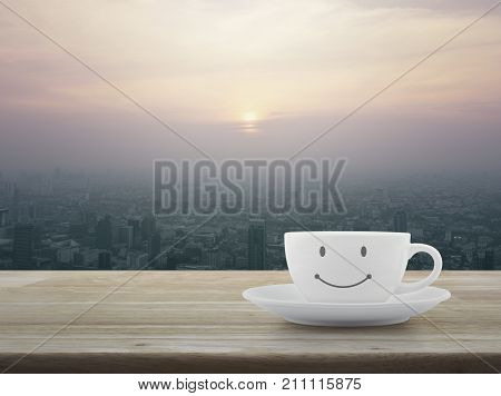 White coffee cup with smile face shape on wooden table over city tower at sunset vintage style Good morning concept