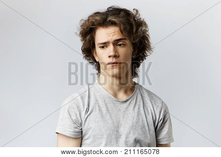 Worried young male with curly hair pondering at blank grey wall. Frustrated 20 year old guy having thoughtful pensive expression on his smooth face worrying about his future life and career