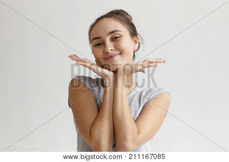 Positive joyful young woman with beautiful features and charming smile placing hands under chin and smiling cheerfully at camera enjoying good day or pleasant compliment. People and happiness concept