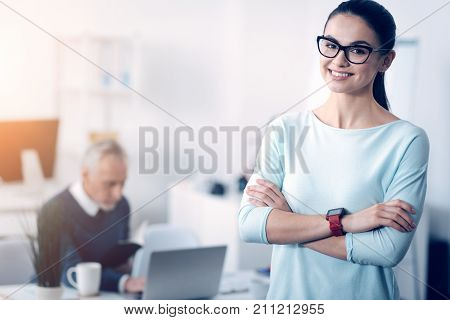Loving my job. Cheerful up and coming specialist in casual looking into the camera with a broad smile on her face while standing in an office with her arms crossed and posing.