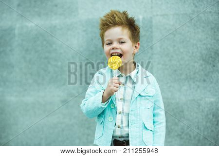 Fashion baby boy in mint jacket and sneakers stands on a gray wall background. Trendy boy with lollipop standing on the street.