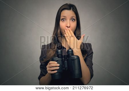 Young woman holding in hands binoculars in front of her and covers her mouth with her hand. Find and search.