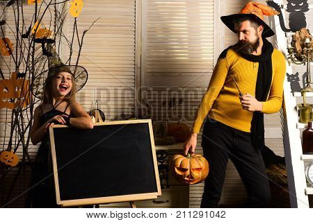 Wizard and little witch hold jack o lantern. Dad and daughter with Halloween decor. Halloween party concept. Girl and bearded man with cheerful faces near blackboard on spooky carnival room background