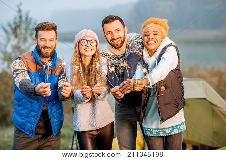 Multi ethnic group of friends dressed in sweaters celebrating with bengal fire standing together at the camping during the evening light