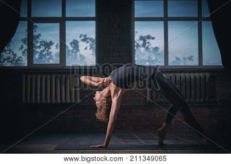 Beautiful sporty fit yogini woman practices yoga asana Wild Thing Pose in the dark hall.