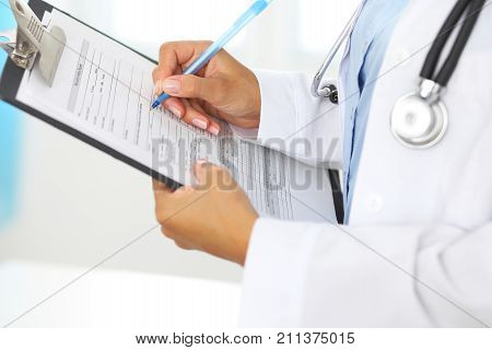 Close-up of a female doctor filling  up medical form at clipboard while standing straight in hospital.
