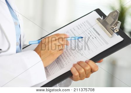 Close-up of a female doctor filling  up medical form at clipboard while standing straight in hospital.
