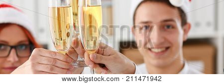 A group of businesspeople celebrating Christmas New Year corporate in the office while toasting holding glasses of champagne in hands on their heads wearing Santa cap.
