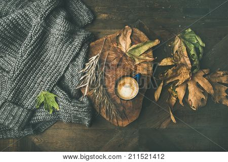 Autumn or Fall morning coffee concept. Flat-lay of knitted woolen grey sweater, wooden tray, mug of coffee and yellow fallen leaves over dark rustic wooden table background, top view