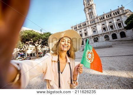 Young woman tourist making selfie photo with portuguese flag in front of the city hall building during the morning light in Porto, Portugal