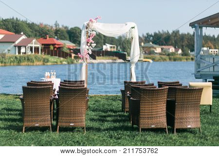 The wedding arc is decorated with flowers at the wedding ceremony. Front view through armchairs. Lake on background.