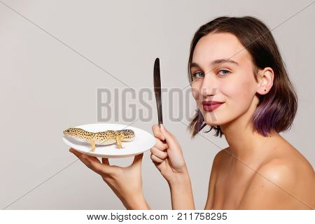 A girl with a knife holds a plate on which a lizard on a gray background and smiles