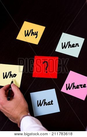 hand with pen pointing on colored notes with w questions black wooden background