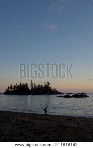 A camper washes a pot in the ocean at sunset Rugged Point Vancouver Is. BC. A cresent moon above and islands off shore complete this beach scene.