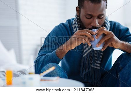 Little cup. Serious concentrated calm ill man sitting quietly while drinking warm tasty tea in his bedroom
