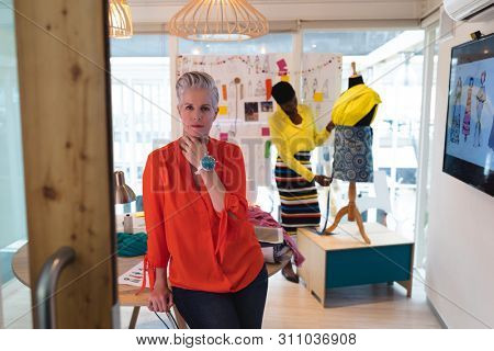 Portrait of Caucasian female fashion designer standing in design studio. African american woman working on dress in the background. This is a casual creative start-up business office for a diverse