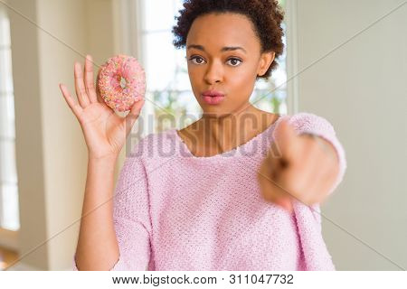 Young african american woman eating pink sugar donut pointing with finger to the camera and to you, hand sign, positive and confident gesture from the front
