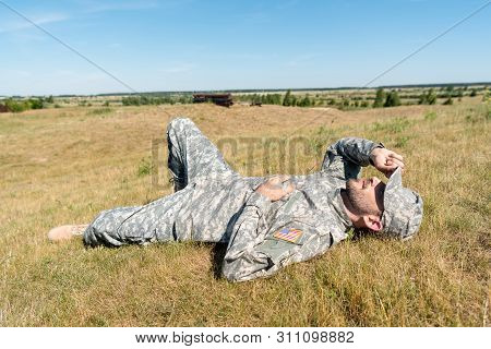 Cheerful Soldier In Military Uniform Lying On Grass And Touching Cap