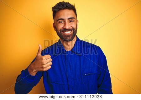 Handsome indian worker man wearing uniform over isolated yellow background doing happy thumbs up gesture with hand. Approving expression looking at the camera with showing success.
