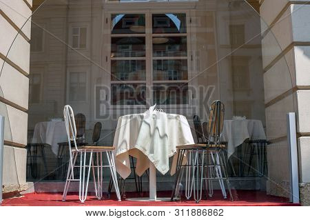 Table Covered With White Tablecloth And Pair Of Chairs On Summer Cafe Terrace Behind Glass Bulkheads
