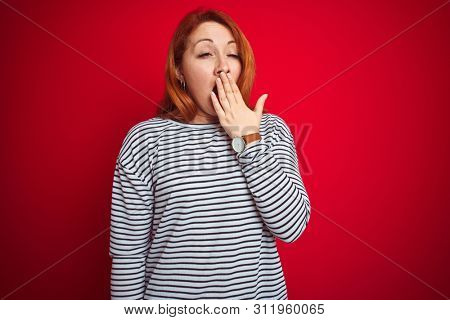 Young redhead woman wearing strapes navy shirt standing over red isolated background bored yawning tired covering mouth with hand. Restless and sleepiness.