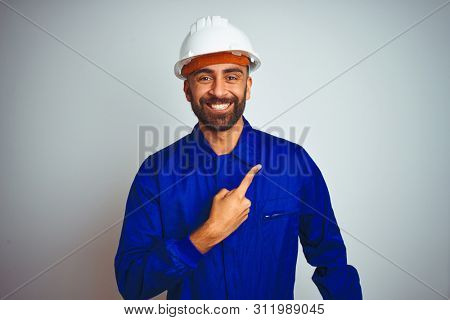 Handsome indian worker man wearing uniform and helmet over isolated white background cheerful with a smile of face pointing with hand and finger up to the side with happy and natural expression 