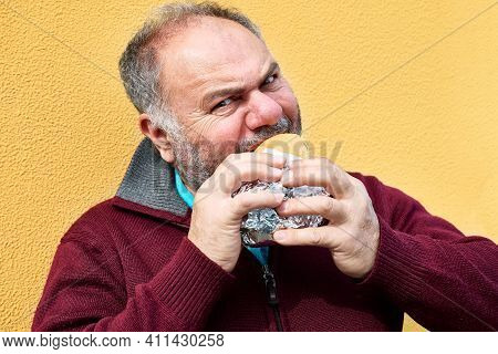Funny Bearded Mature Man Eating Sandwich On The Street Against A Yellow Wall. Man Hungry Like A Wolf