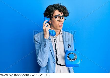 Hispanic young man speaking on vintage telephone in shock face, looking skeptical and sarcastic, surprised with open mouth 