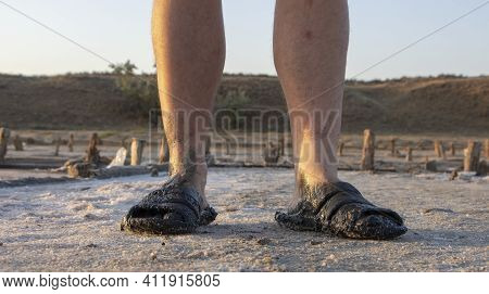 Men's Bare Feet And Summer Shoes Are Dirty In The Swamp, The Salt Of The Salt Lake Underfoot. The Sh