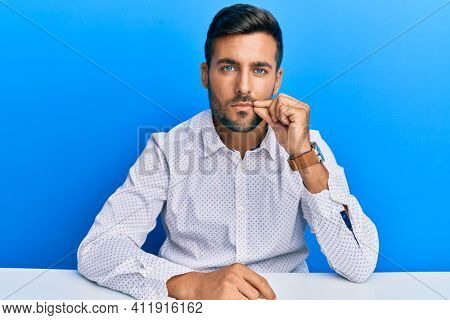 Handsome hispanic man wearing business clothes sitting on the table mouth and lips shut as zip with fingers. secret and silent, taboo talking 