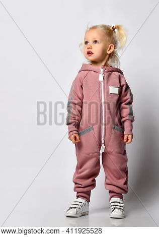 Little Preschooler Girl In Warm Pink Sportswear On A Gray Background. Child Looks To The Side, His H