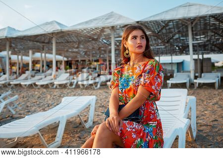 A Young Tanned Woman In A Red Dress, Posing Sitting On A Chaise Longue With Her Arms Crossed. Sandy 