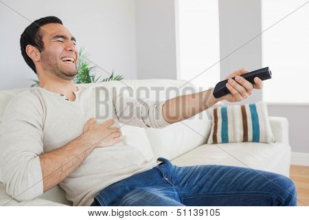 Handsome man bursting out laughing while watching tv on cosy sofa