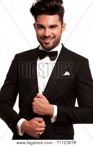 smiling young groom in tuxedo and bowtie holding his suit on white background