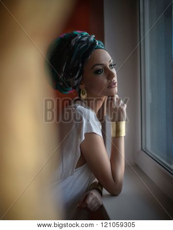 Attractive sexy lady in white blouse posing in window frame looking outside. Portrait of sensual young woman with turban. Gorgeous female with beautiful eyes daydreaming enjoying the bright day light